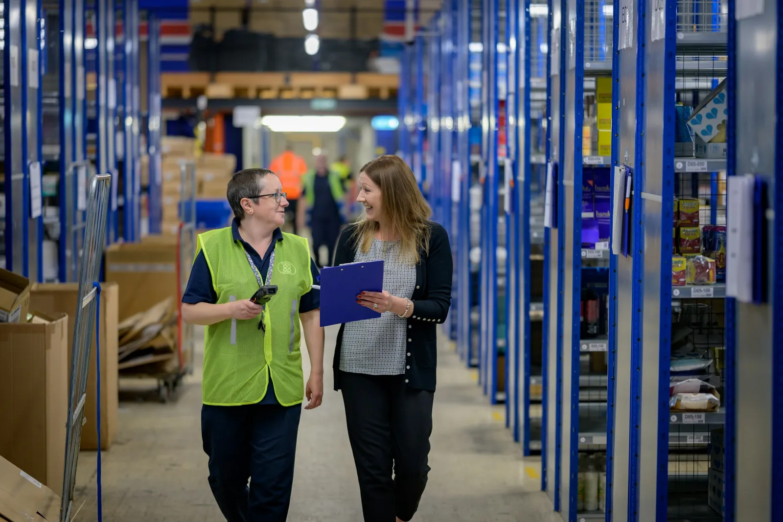 Two warehouse staff reviewing stock while walking through storage aisles